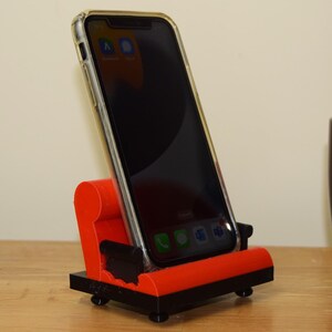 May include: A red and black phone stand with a smartphone in a clear case. The phone is displaying several apps on the screen. The stand is on a wooden surface.