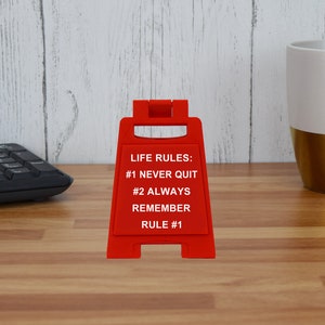May include: A red sign with the text "LIFE RULES: #1 NEVER QUIT #2 ALWAYS REMEMBER RULE #1" stands on a wooden desk. A black keyboard and a white and gold mug are in the background.