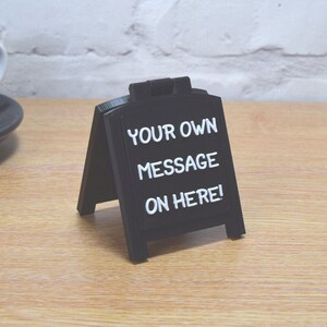 May include: A black A-frame chalkboard sign with white text reading "YOUR OWN MESSAGE ON HERE!". The sign is displayed on a wooden surface. A coffee cup and saucer are visible in the background. The sign is designed for custom messages.