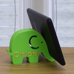 May include: Green elephant-shaped phone stand with the name "Chloe" printed on the side. A black tablet is propped up in the stand.