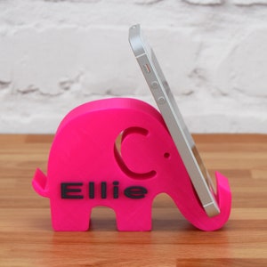 May include: A pink 3D printed elephant phone stand with the name "Ellie" printed on the side. A silver smartphone is propped up in the stand.