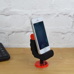 May include: A 3D printed phone stand shaped like a person sitting in a chair. The stand is black with red accents and the word "Here" is printed on the base. A white smartphone is in the stand.