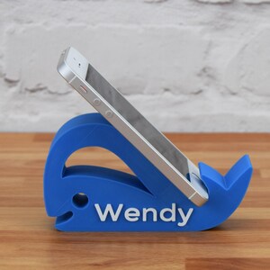 May include: A blue whale-shaped phone stand with the name "Wendy" printed on it. A white smartphone is resting in the stand.