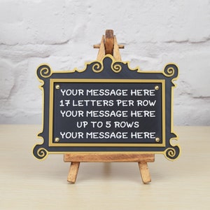 May include: A black chalkboard sign with gold trim and a wooden easel. The sign has the text "YOUR MESSAGE HERE 17 LETTERS PER ROW YOUR MESSAGE HERE UP TO 5 ROWS YOUR MESSAGE HERE" written in white.