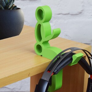 May include: A green 3D printed headphone stand shaped like a person sitting down. The stand has a hook on the back for hanging headphones. The stand is on a wooden table.