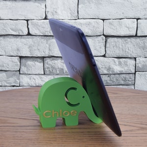 Pode incluir: Um suporte de telefone verde em forma de elefante com o nome "Chloe" gravado nele. Um tablet preto est&aacute; apoiado no suporte.