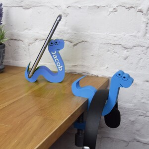 May include: Two blue snake-shaped phone and headphone holders. The phone holder has the name "Jacob" written on it. The headphone holder is attached to a desk with a clamp.
