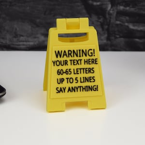 May include: A yellow plastic warning sign with black text. The sign reads "WARNING! YOUR TEXT HERE 60-65 LETTERS UP TO 5 LINES SAY ANYTHING!" The sign is in an A-frame shape, with a hinged top.