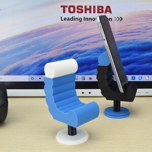 May include: Two phone stands, one shaped like a chair and the other shaped like a hand, both in blue and white. The chair stand has a white base and the hand stand has a blue base. Both stands are on a wooden surface.