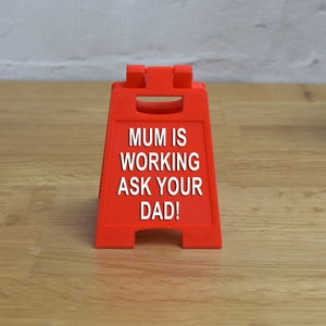 May include: A red, triangular sign with the text "MUM IS WORKING ASK YOUR DAD!" printed in white.