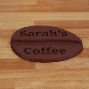 May include: A brown ceramic coffee cup coaster shaped like a coffee bean with the text "Sarah's Coffee" engraved on it.