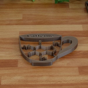 May include: A brown, cup-shaped coaster with the word "Caffeine" and a molecular structure design. The coaster is next to a white coffee cup and a decorative white wooden box with a heart-shaped cutout.