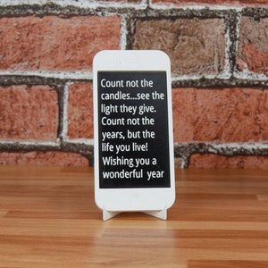 May include: A white smartphone stand with a black screen displaying a quote about counting the life you live, not the years. The quote reads "Count not the candles...see the light they give. Count not the years, but the life you live! Wishing you a wonderful year."