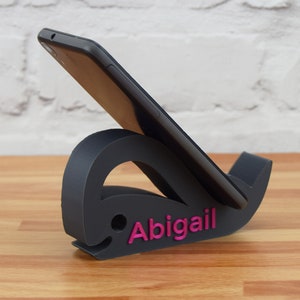 May include: A black phone stand shaped like a whale with the name "Abigail" printed in pink on the side.