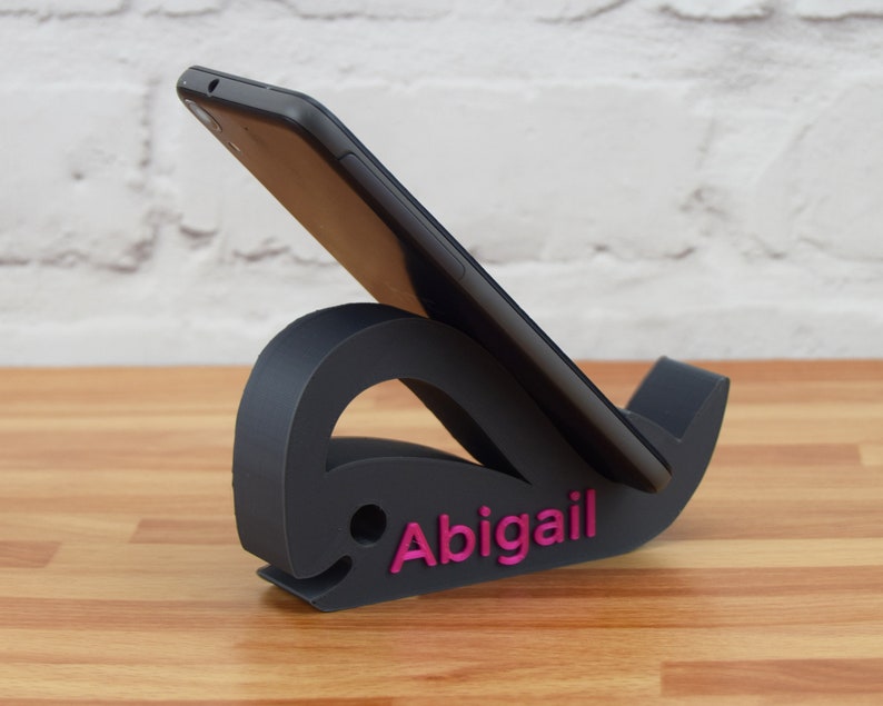 May include: A black phone stand shaped like a whale with the name "Abigail" printed in pink on the side.