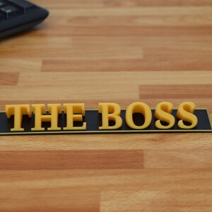 May include: A black plaque with a gold border and the words "THE BOSS" in yellow 3D letters.