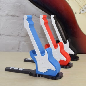 Puede incluir: Cuatro soportes de guitarra en miniatura en azul, rojo, negro y blanco. Los soportes est&aacute;n hechos de pl&aacute;stico y tienen forma de guitarras el&eacute;ctricas. Los soportes est&aacute;n dispuestos en fila de izquierda a derecha, con el soporte azul en la parte delantera.