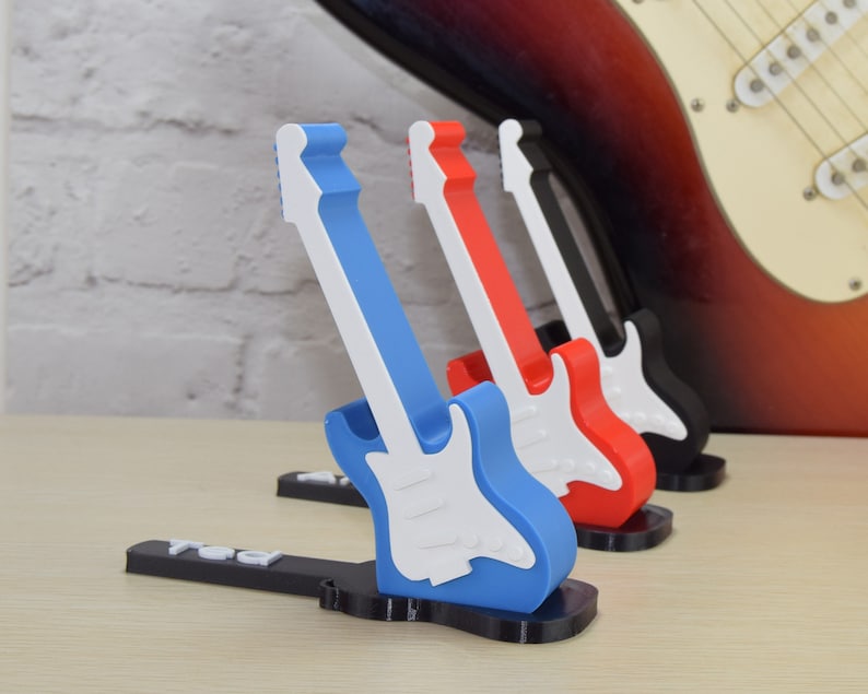 Puede incluir: Cuatro soportes de guitarra en miniatura en azul, rojo, negro y blanco. Los soportes est&aacute;n hechos de pl&aacute;stico y tienen forma de guitarras el&eacute;ctricas. Los soportes est&aacute;n dispuestos en fila de izquierda a derecha, con el soporte azul en la parte delantera.