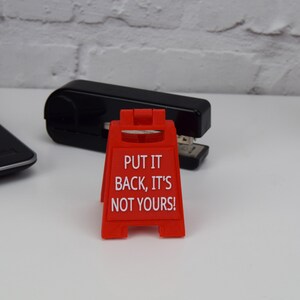 May include: A red plastic sign with the text "PUT IT BACK, IT'S NOT YOURS!" stands in front of a black stapler. The sign is a miniature version of a caution sign. A laptop is partially visible in the background.