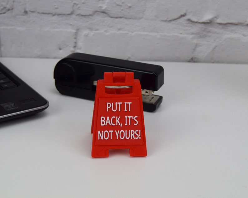 May include: A red plastic sign with the text "PUT IT BACK, IT'S NOT YOURS!" stands in front of a black stapler. The sign is a miniature version of a caution sign. A laptop is partially visible in the background.
