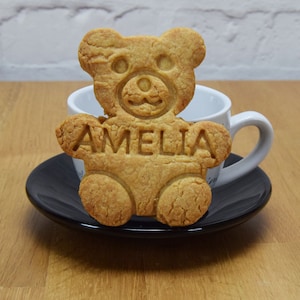 Puede incluir: Una galleta de az&uacute;car moreno con forma de osito de peluche con el nombre Amelia escrito en ella. La galleta est&aacute; sobre una taza de t&eacute; y un platillo blancos.