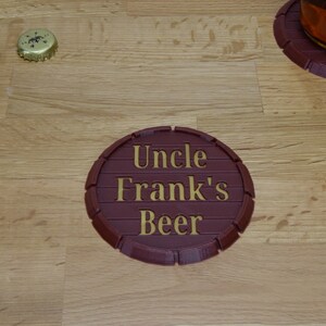 May include: A brown, round, wooden coaster with a raised edge. The coaster has the words "Uncle Frank's Beer" written in gold lettering.