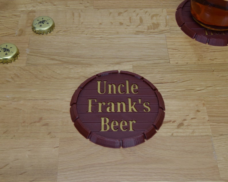 May include: A brown, round, wooden coaster with a raised edge. The coaster has the words "Uncle Frank's Beer" written in gold lettering.