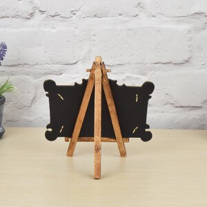 May include: A small wooden easel with a black chalkboard frame. The easel is made of natural wood and has a rustic look. The chalkboard is blank and ready to be written on.