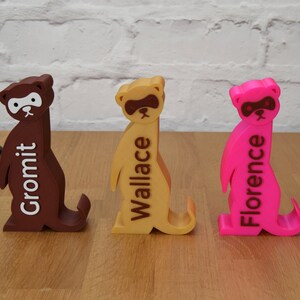 May include: Three 3D printed meerkat figurines, each painted a different color: brown, yellow, and pink. The meerkats are standing upright with their arms raised. Each meerkat has a name printed on its back: Gromit, Wallace, and Florence.