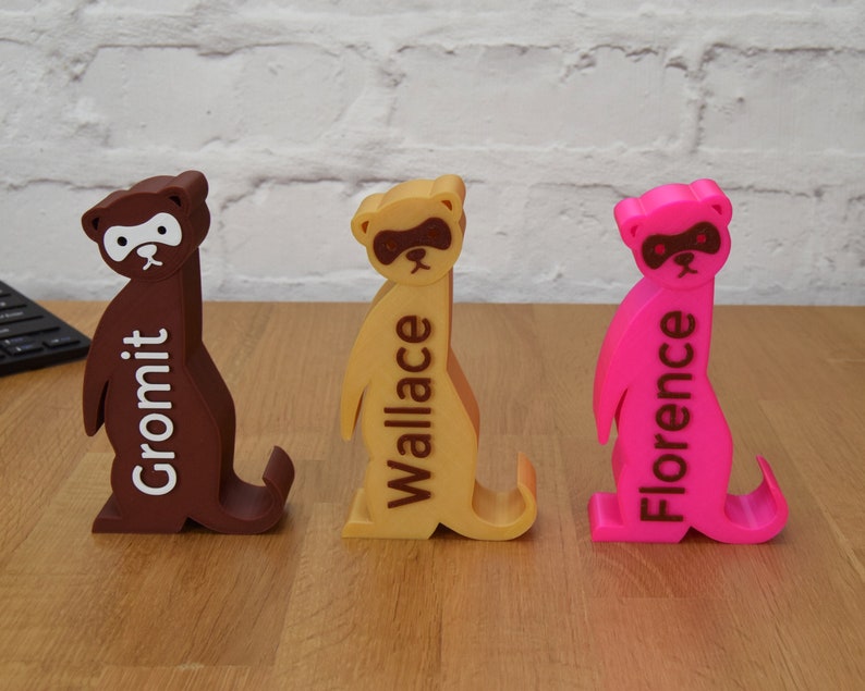 May include: Three 3D printed meerkat figurines, each painted a different color: brown, yellow, and pink. The meerkats are standing upright with their arms raised. Each meerkat has a name printed on its back: Gromit, Wallace, and Florence.
