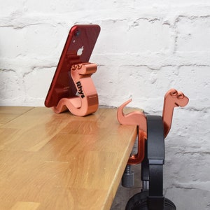 May include: Two rose gold colored phone and headphone holders shaped like dinosaurs. The phone holder is holding a red iPhone. The headphone holder is attached to a desk with a clamp and has a black headphone hanging from it.