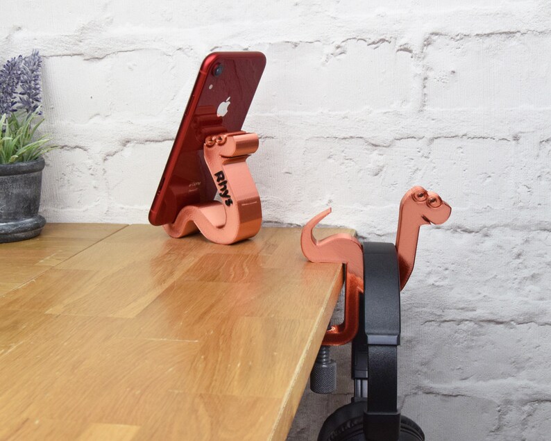 May include: Two rose gold colored phone and headphone holders shaped like dinosaurs. The phone holder is holding a red iPhone. The headphone holder is attached to a desk with a clamp and has a black headphone hanging from it.
