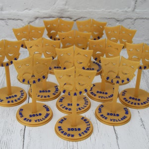 May include: A collection of 15 gold-colored plastic trophies with a comedy and tragedy mask design. Each trophy has a round base with blue text that reads "ELAPS BIG VILLAGE" and the year "2019". The text on the bases includes the names of different groups, such as "The Deewives", "The Meadow Strollers", and "The Outzerables".
