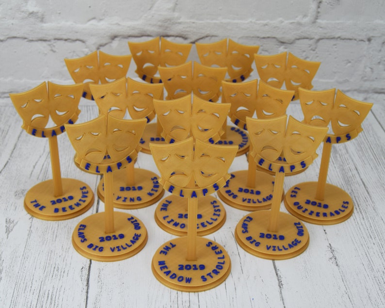May include: A collection of 15 gold-colored plastic trophies with a comedy and tragedy mask design. Each trophy has a round base with blue text that reads "ELAPS BIG VILLAGE" and the year "2019". The text on the bases includes the names of different groups, such as "The Deewives", "The Meadow Strollers", and "The Outzerables".