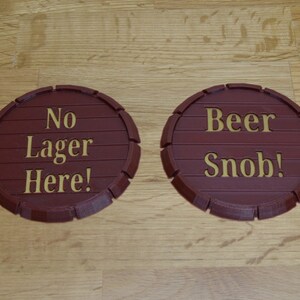 May include: Two brown, barrel-shaped coasters with gold lettering. One reads "No Lager Here!" and the other says "Beer Snob!" The coasters are made of a ridged material, resembling a wooden barrel.