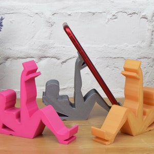 May include: Three 3D printed phone stands in the shape of people, one pink, one gray, and one orange. The stands are designed to hold a phone upright.