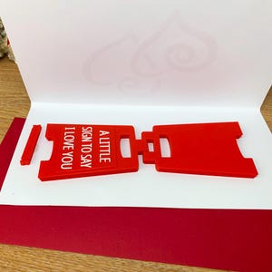 Op de afbeelding: Een rood, plastic bord met de tekst "A little sign to say I love you". Het bord is dubbelgevouwen en ligt op een wit oppervlak.