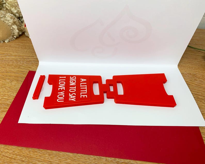 Op de afbeelding: Een rood, plastic bord met de tekst "A little sign to say I love you". Het bord is dubbelgevouwen en ligt op een wit oppervlak.