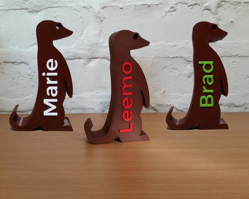 K&ouml;nnte beinhalten: Drei braune Telefonhalter in Form eines Erdm&auml;nnchens mit den Namen "Marie", "Leemo" und "Brad", die in wei&szlig;en, roten und gr&uuml;nen Buchstaben darauf geschrieben sind.