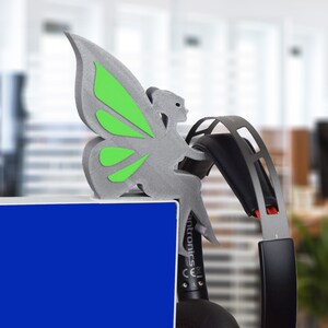K&ouml;nnte beinhalten: Eine graue, 3D-gedruckte Schmetterlingsfigur mit gr&uuml;nen Fl&uuml;geln, die auf einer blauen Oberfl&auml;che sitzt. Der Schmetterling sitzt auf einem schwarzen und roten Headset.