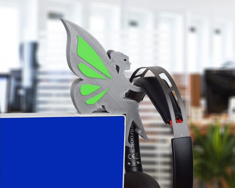 K&ouml;nnte beinhalten: Eine graue, 3D-gedruckte Schmetterlingsfigur mit gr&uuml;nen Fl&uuml;geln, die auf einer blauen Oberfl&auml;che sitzt. Der Schmetterling sitzt auf einem schwarzen und roten Headset.
