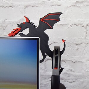 May include: A black dragon-shaped headphone stand with red accents. The dragon is perched on the edge of a computer monitor, with its wings and tail extending down. The dragon is spewing red flames from its mouth. A headset is hanging from the stand.