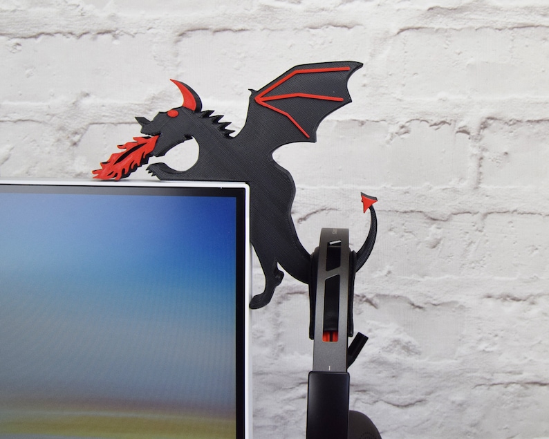 May include: A black dragon-shaped headphone stand with red accents. The dragon is perched on the edge of a computer monitor, with its wings and tail extending down. The dragon is spewing red flames from its mouth. A headset is hanging from the stand.