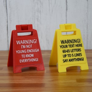 May include: Two plastic warning signs, one red and one yellow. The red sign reads "WARNING! I'M NOT YOUNG ENOUGH TO KNOW EVERYTHING!" The yellow sign says "WARNING! YOUR TEXT HERE 60-65 LETTERS UP TO 5 LINES SAY ANYTHING!"