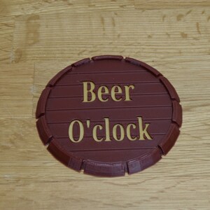 May include: A round, brown coaster with the words "Beer O'clock" in gold lettering. The coaster is designed to resemble a barrel end, with horizontal lines and a curved edge.