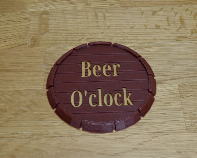 May include: A round, brown coaster with the words "Beer O'clock" in gold lettering. The coaster is designed to resemble a barrel end, with horizontal lines and a curved edge.