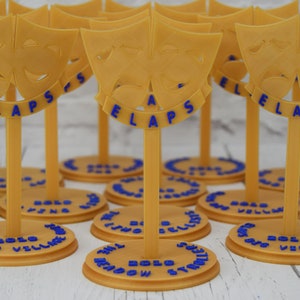 May include: A set of gold-colored, 3D printed, table-top awards with a blue inscription that reads "The Meadow Synodic 2020 Village Bells". Each award features a pair of stylized masks, one smiling and one frowning.