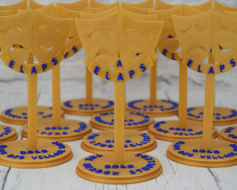 May include: A set of gold-colored, 3D printed, table-top awards with a blue inscription that reads "The Meadow Synodic 2020 Village Bells". Each award features a pair of stylized masks, one smiling and one frowning.