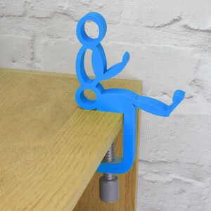 May include: A blue plastic figurine of a person sitting on a clamp. The clamp is attached to a wooden surface.