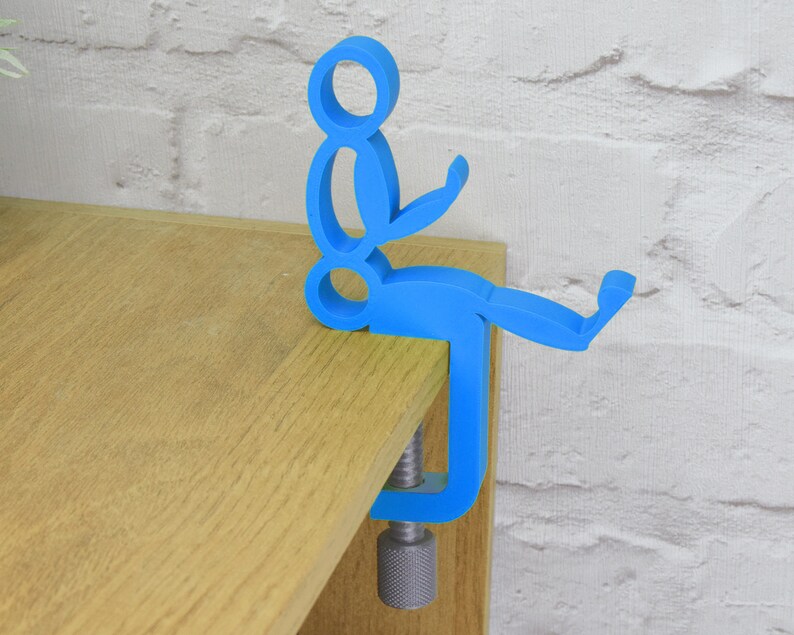 May include: A blue plastic figurine of a person sitting on a clamp. The clamp is attached to a wooden surface.